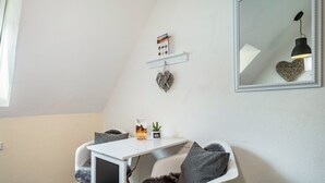 Quarto moderno com escrivaninha branca, cadeira com almofadas cinzentas, uma pequena planta em vaso, espelho emoldurado e um ornamento em formato de coração na parede.