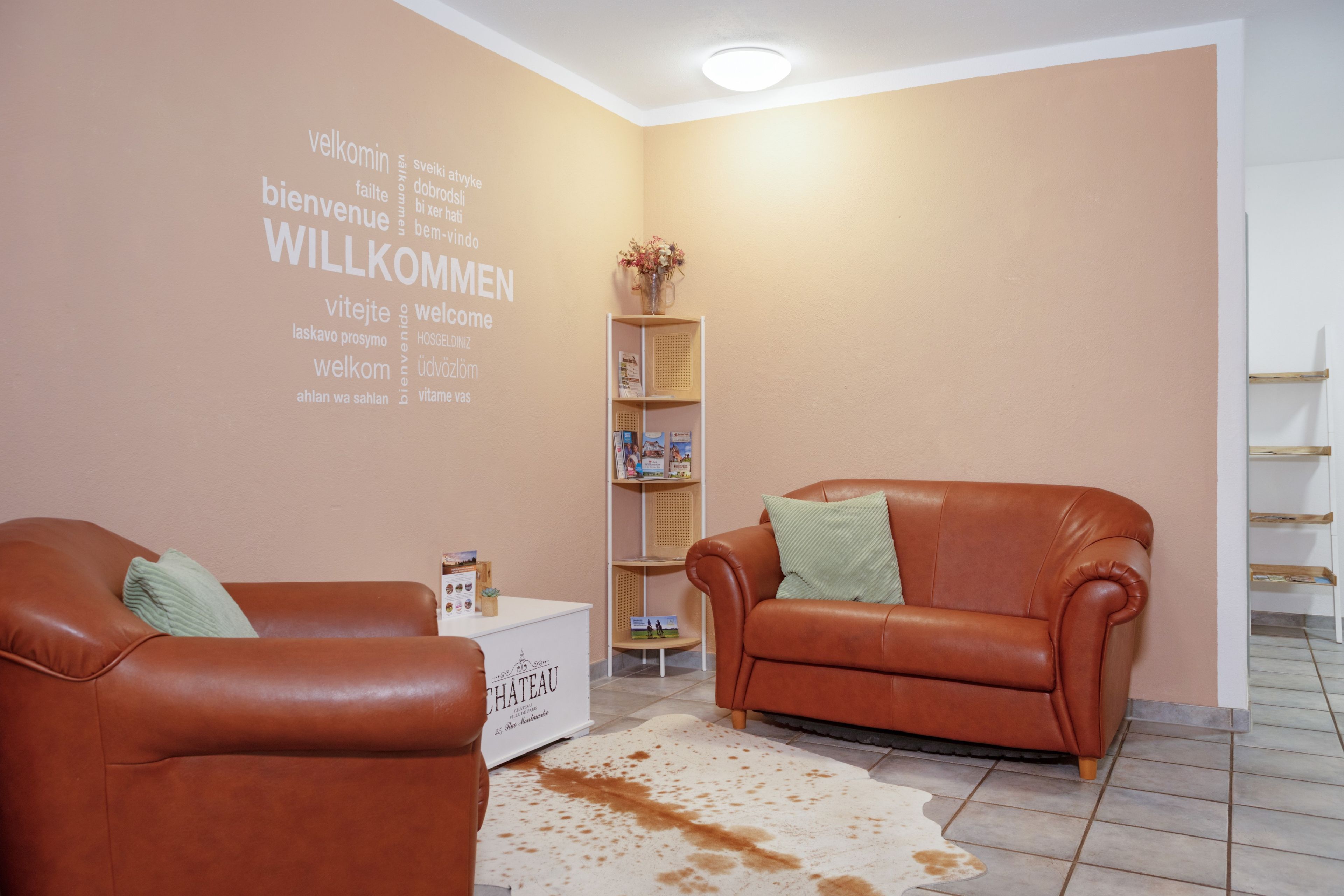 Ein Zimmer mit zwei Ledersofas, einem Couchtisch, einem Bücherregal und einer Wand mit mehrsprachigem Willkommenstext.