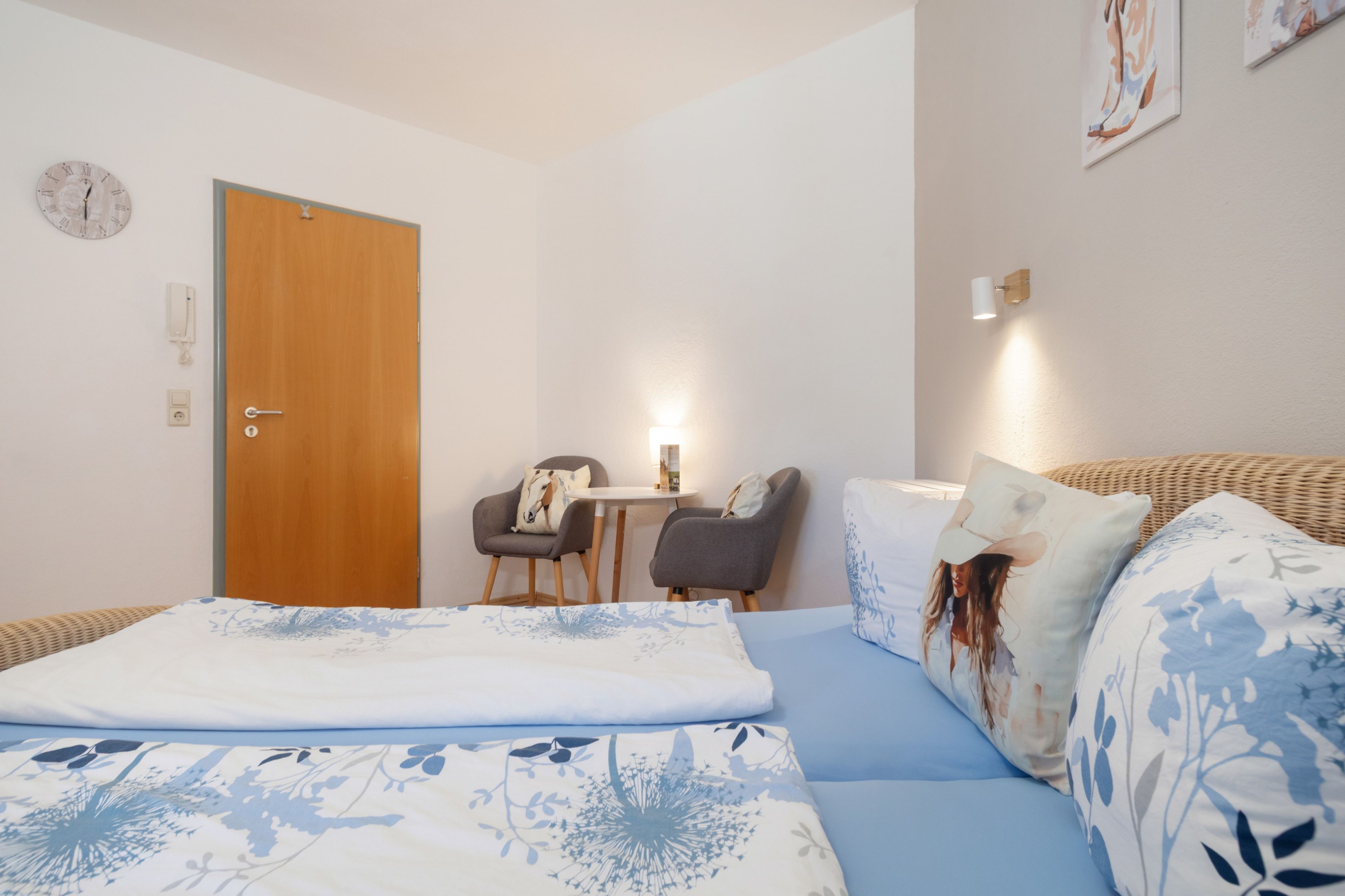 Ein Schlafzimmer mit einem Bett, einer Holztür, einem kleinen Tisch mit Stühlen und einer Wanduhr.