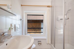 Ein Badezimmer mit einem weißen Waschbecken, einem Spiegel, einer Dusche und einem Fenster mit einem Holzrollo.