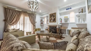 Un salon avec un canapé, une table basse et une cheminée. On y trouve des rideaux, un lustre et des étagères ornées d’objets décoratifs.