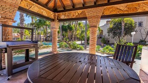 Un espace extérieur abrité, doté d’une table et de chaises en bois, entouré de palmiers et bordé d’une piscine.