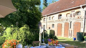 Ein Essbereich im Freien mit einem Tisch für zwei Personen, umgeben von Grünpflanzen und einem Gebäude mit einem Ziegeldach.