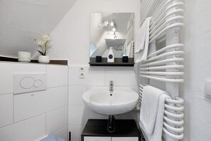 Un baño moderno con un lavabo blanco, un espejo, un perchero para toallas y un pequeño estante con una flor y una botella.
