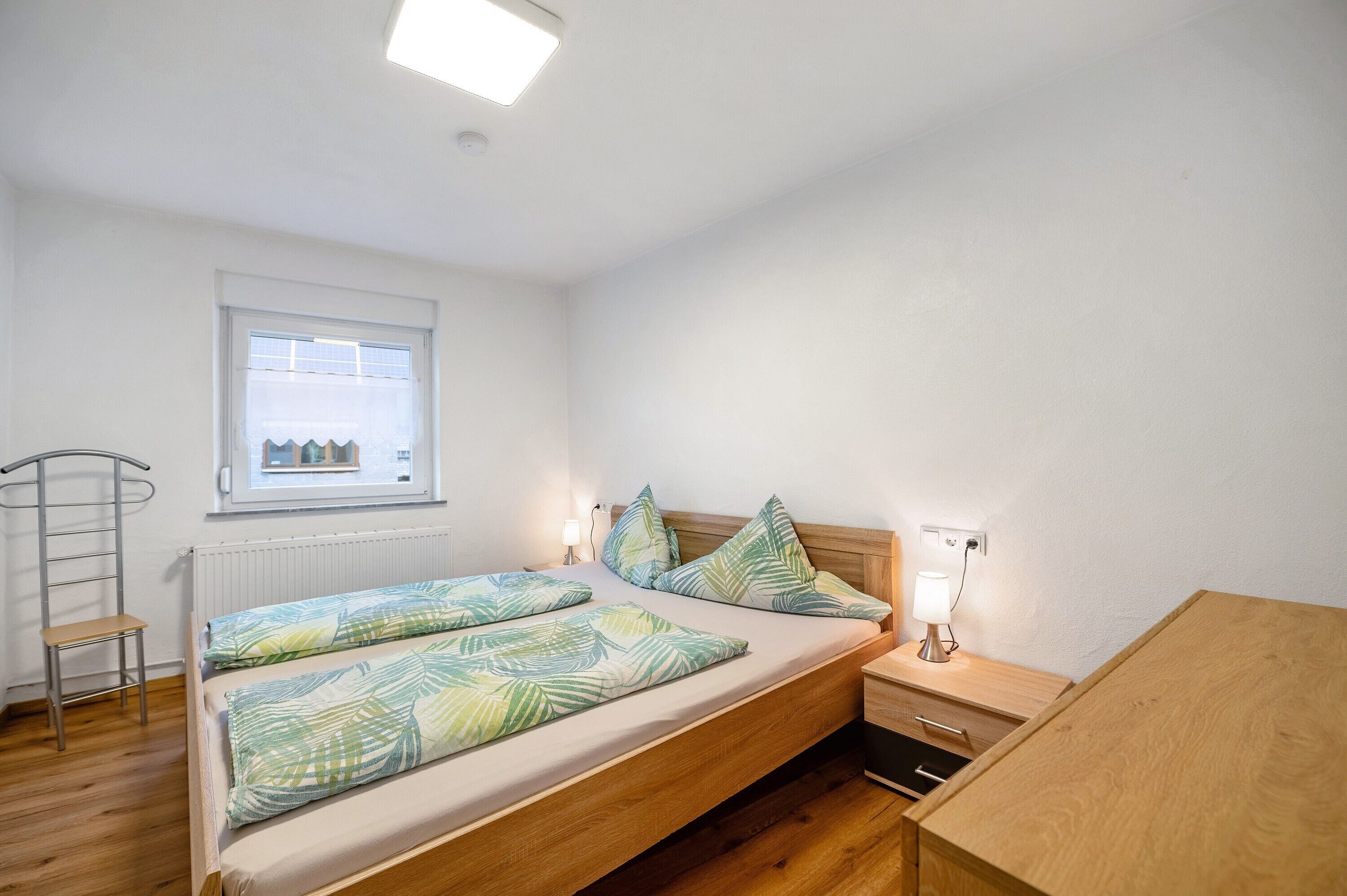 Een slaapkamer met een bed, nachtkastje, lamp, raam, radiator en een houten stoel.