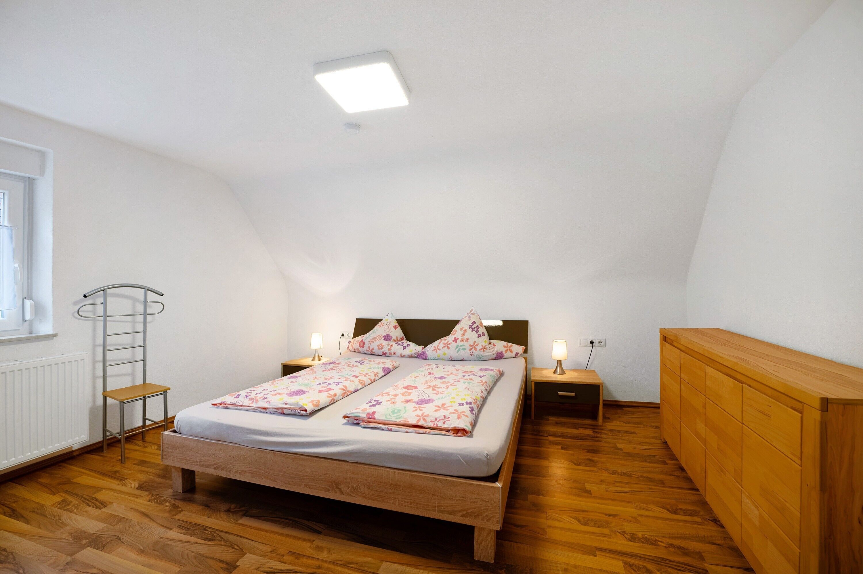 A bedroom with a wooden bed, a nightstand, a dresser, a radiator, and a window.