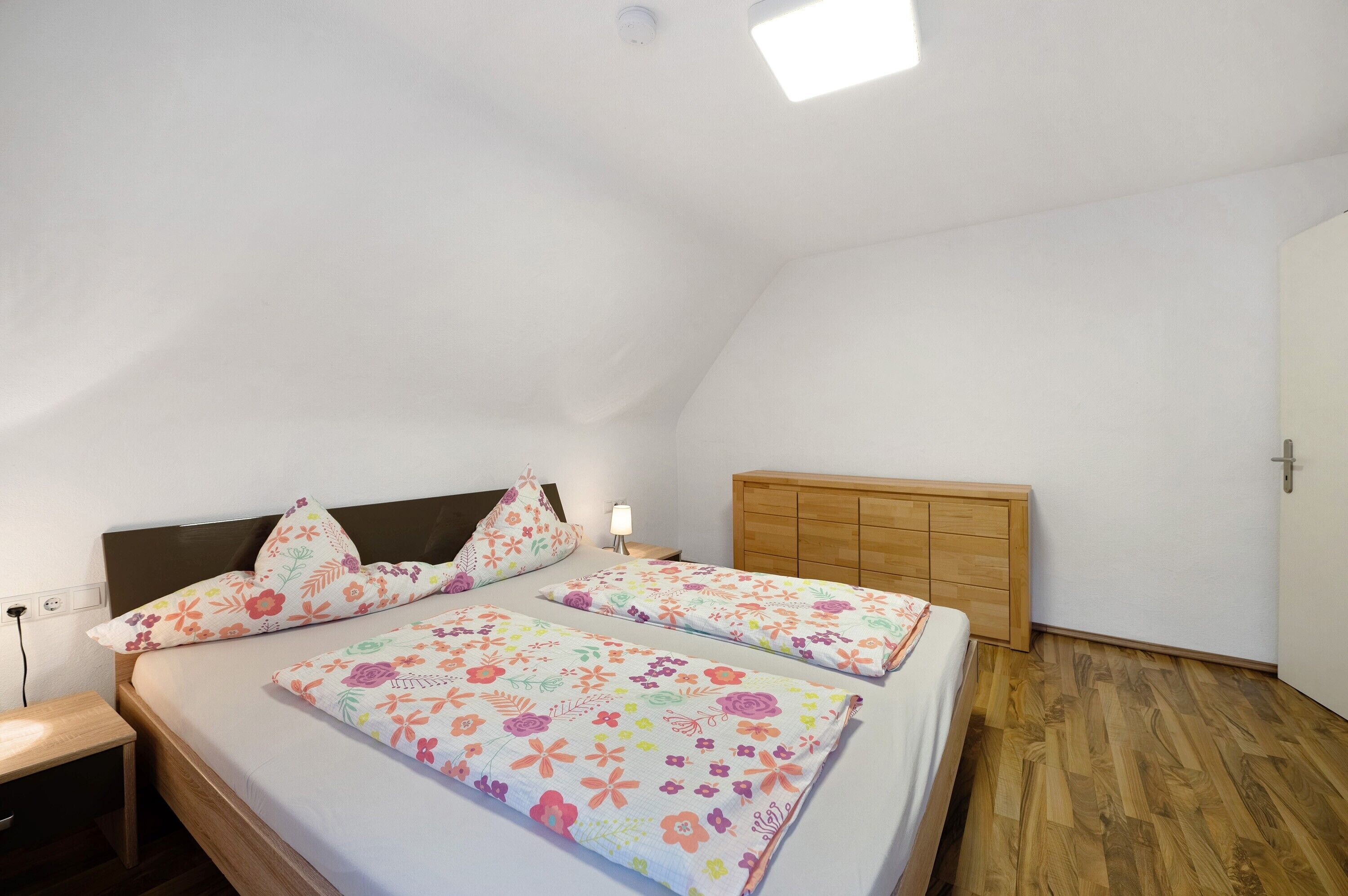 A bedroom with two beds, a nightstand, a wooden wardrobe, and a ceiling light.