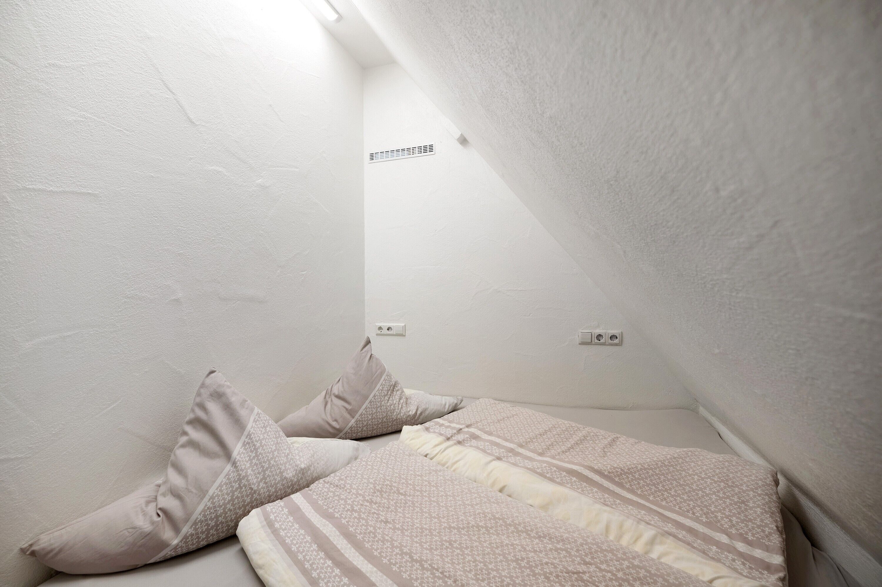 A small, white-walled room with a sloped ceiling, a bed with patterned bedding, and a vent on the wall.