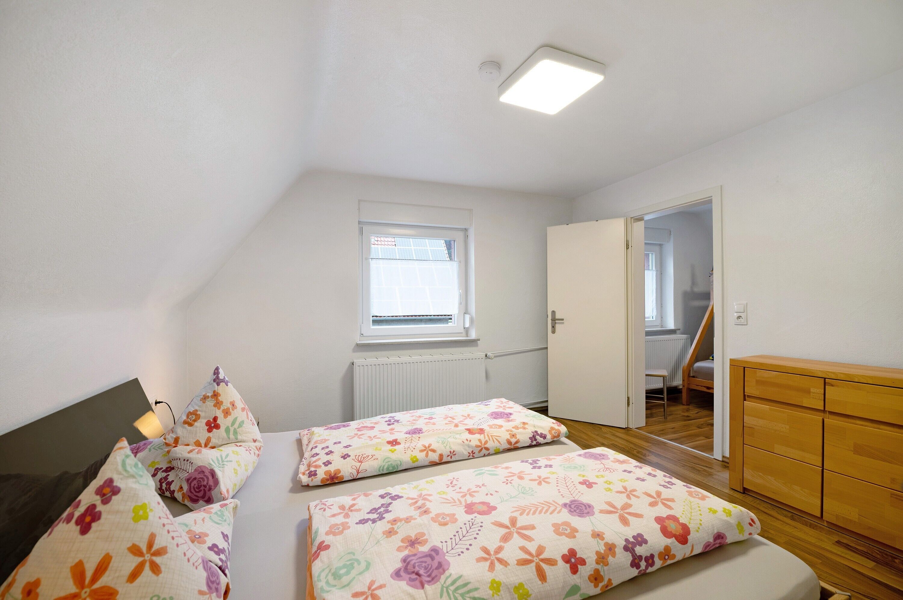 A bedroom with a sloped ceiling, a window, a radiator, a wooden dresser, and a door leading to another room.