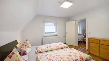 A bedroom with a sloped ceiling, a window, a radiator, a wooden dresser, and a door leading to another room.