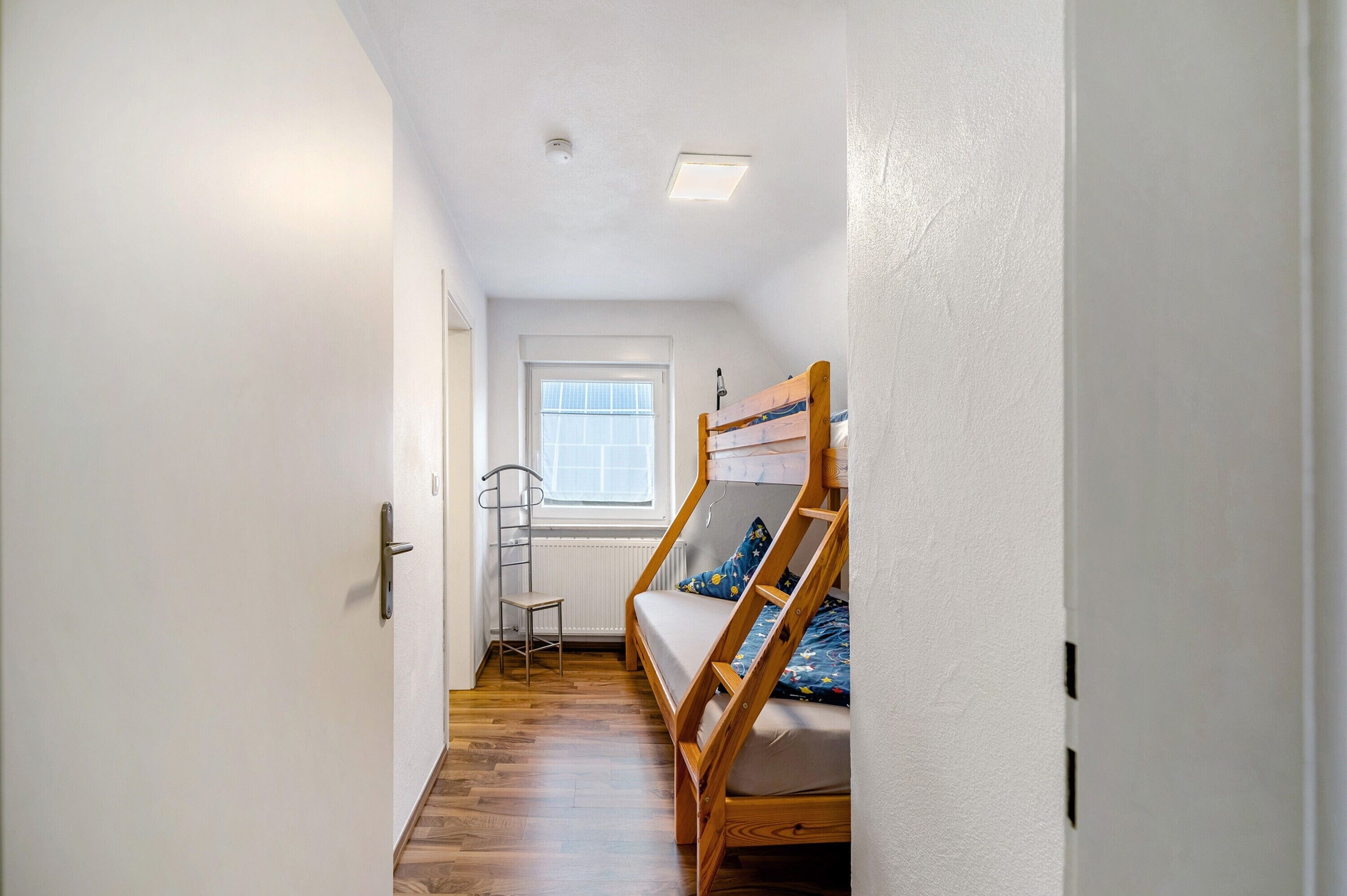 Ein schmaler Flur, der zu einem Schlafzimmer mit Etagenbett, einem Fenster mit Jalousien, einer Heizung und einer Tür führt.