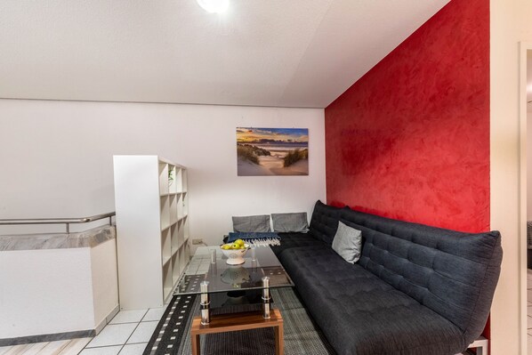 A modern living area with a glass coffee table, a dark sofa, a red accent wall, and a painting of a beach scene.