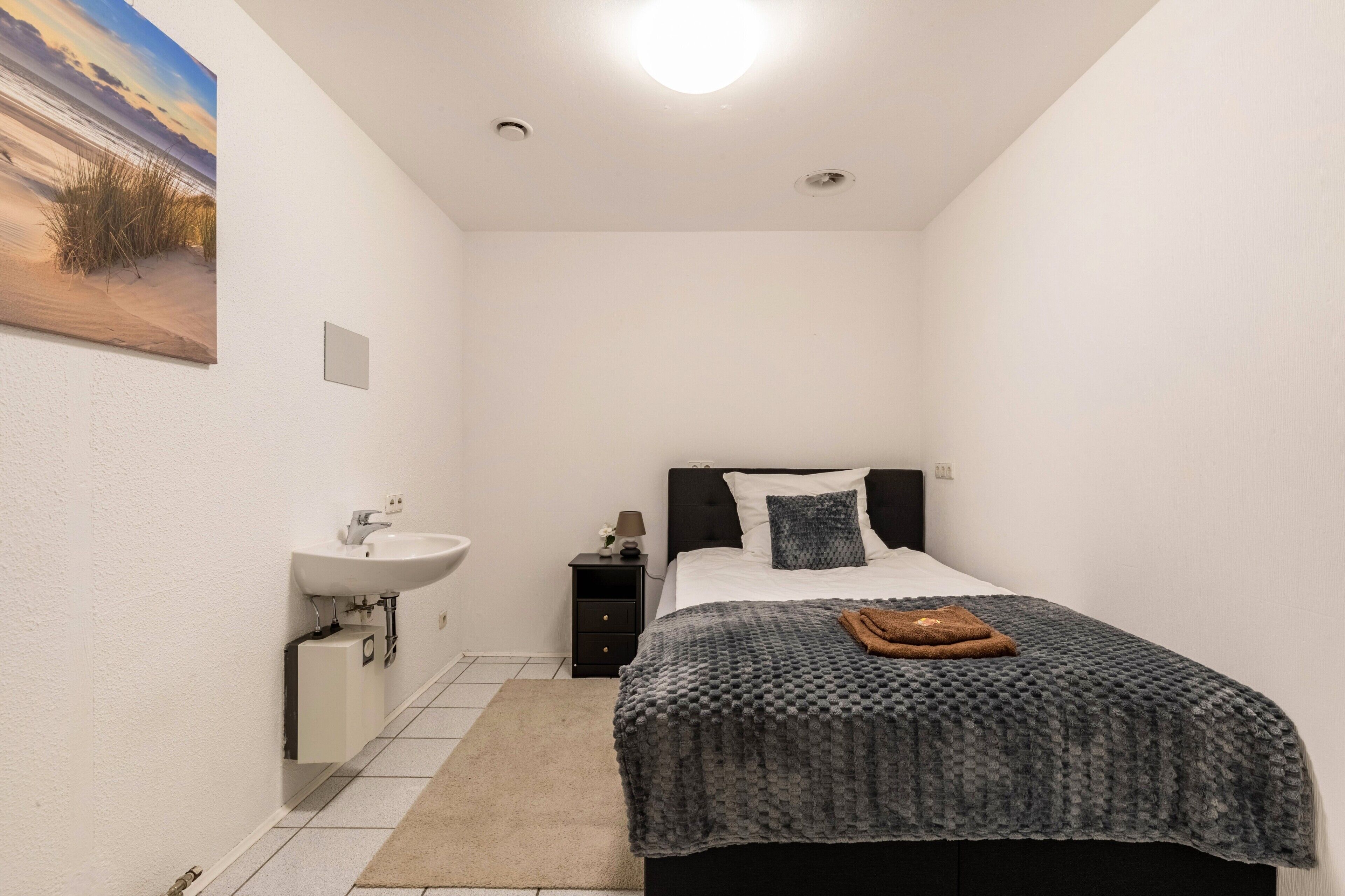A small, modern bedroom with a bed, a bedside table, a nightstand, a wall-mounted sink, and a painting of a beach scene.
