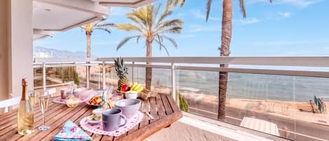 A wooden table set with a bottle, glasses, and fruits, overlooking a coastal view with palm trees.