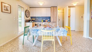 A dining area with a table set for four, chairs, a kitchen in the background, and a door leading to another room. - Rives d'Azur Apt with Sea View Terrace, Menton, France (Menton)