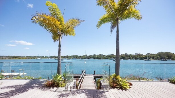 A wooden deck with a glass railing overlooking a marina with boats. - Riverside Elegance on Noosa Parade (Noosa)