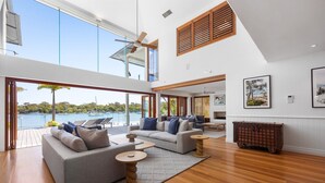 A spacious living area with a large window overlooking a body of water, a ceiling fan, and a view of the outdoors. - Riverside Elegance on Noosa Parade (Noosa)