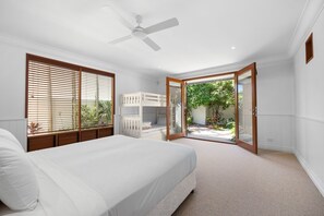 A bedroom with a bed, a ceiling fan, a window with blinds, and an open door leading to a balcony with plants. - Riverside Elegance on Noosa Parade (Noosa)