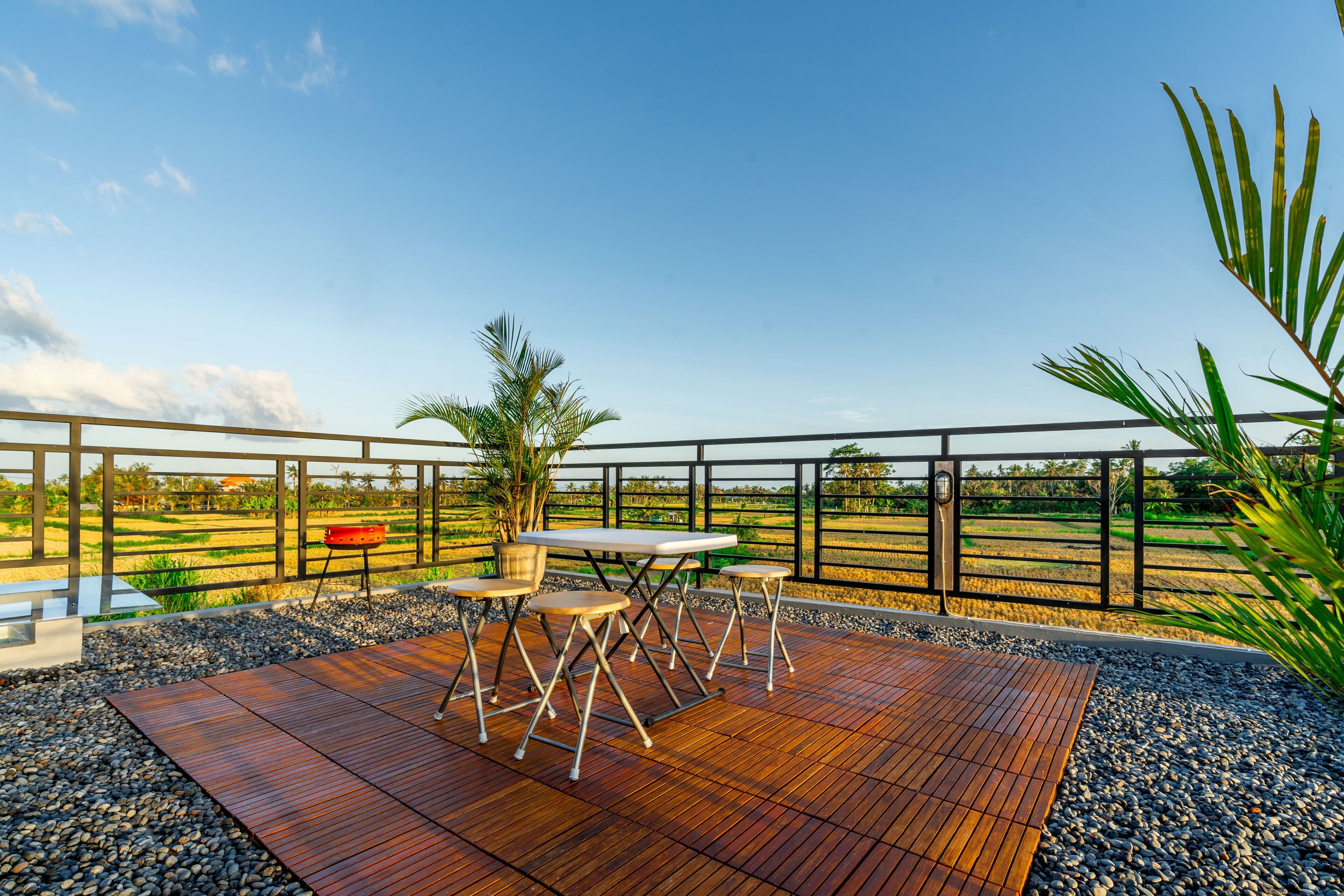 An outdoor deck with a wooden table and chairs, surrounded by a metal railing, overlooking a farm landscape.