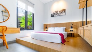 A modern hotel room with a large bed, a wooden headboard, a desk with a chair, and a large window offering a view of greenery. - Feels Like Home Villa (Blahbatuh)