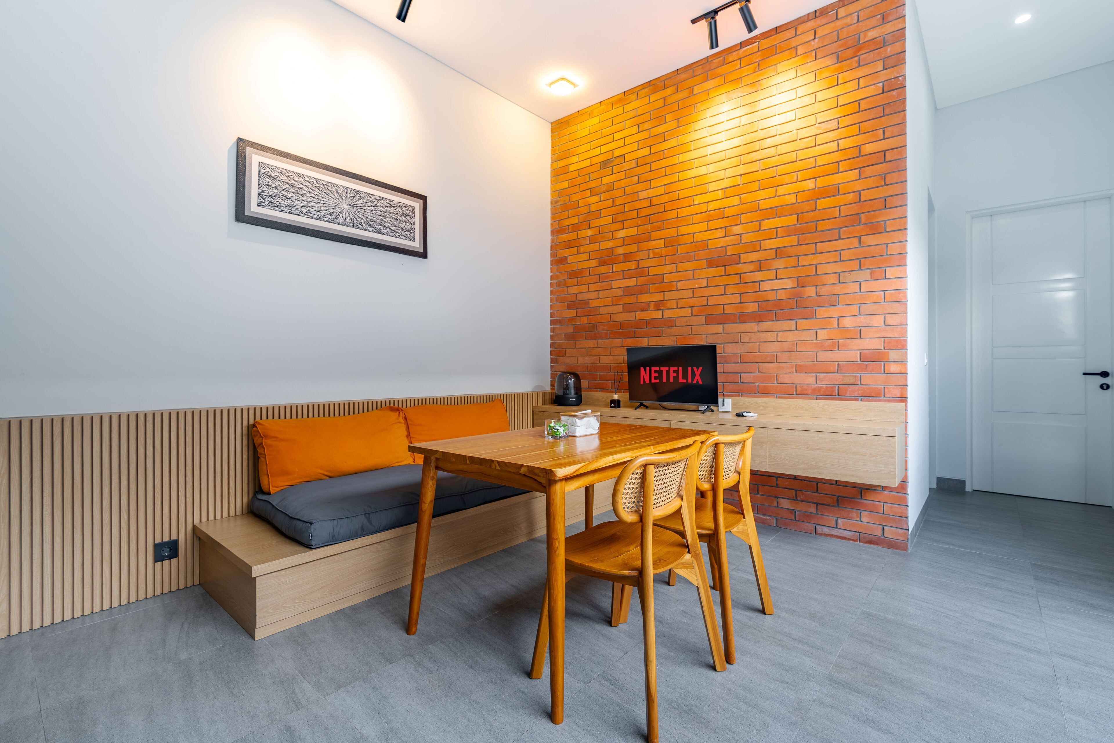 A modern room with a wooden dining table, orange cushions, a brick accent wall, and a Netflix sign.