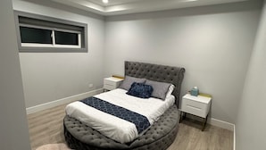 A bedroom with a tufted headboard, a bed with a white and dark blue bedspread, a nightstand with a lamp, and a window with blinds. - Exquisite Chicago Modern Rooftop, Sleeps 9 (Chicago)