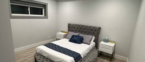 Une chambre à coucher avec une tête de lit capitonnée, un lit recouvert d’une couvre-lit blanche et bleu foncé, une table de chevet avec une lampe et une fenêtre avec des stores.