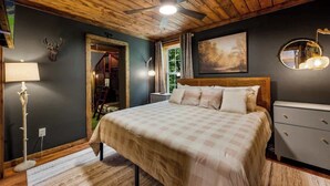 A bedroom with a wooden ceiling, a bed with a checkered bedspread, a nightstand, a bedside lamp, a dresser, and a window with curtains. - *Central! Game Room! Hot Tub!* (Pigeon Forge)