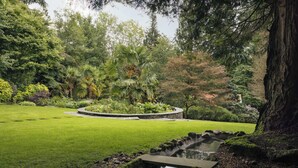 A lush garden with a central circular stone feature, a small water stream, and a variety of trees and shrubs. - High Cross Lodge | Ridgestone (Troutbeck)