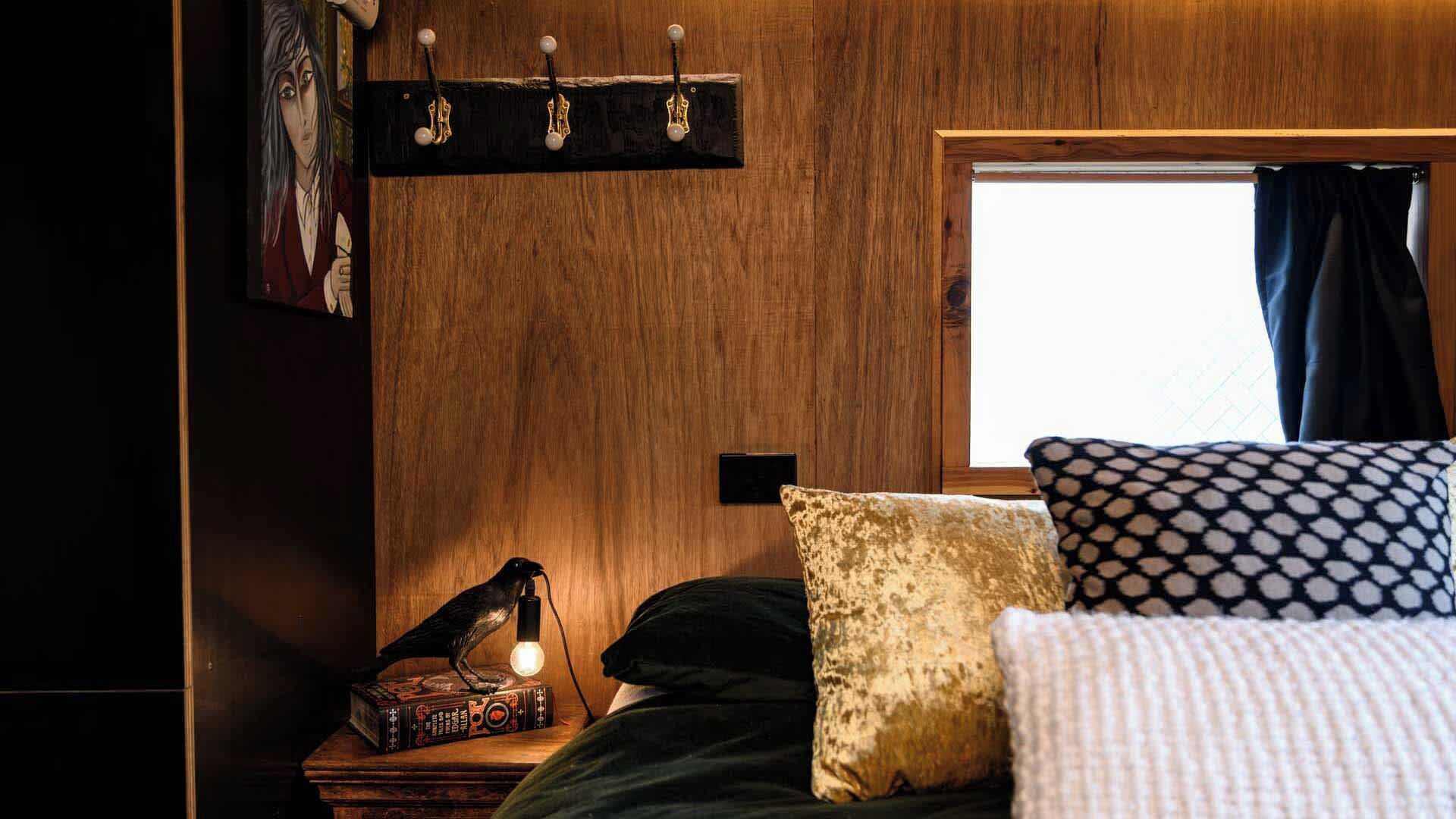 A cozy room with a wooden wall, a bed with patterned pillows, a nightstand with a lamp and a book, and a window with curtains.