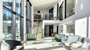 A spacious living area with a modern design, featuring a large window, a staircase, and a seating area with green upholstered sofas.