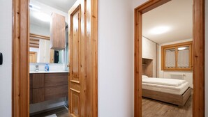 A bedroom with a wooden bed, a window with curtains, and a bathroom with a mirror, sink, and cabinets. - New apartment in the center of Canazei (Canazei)