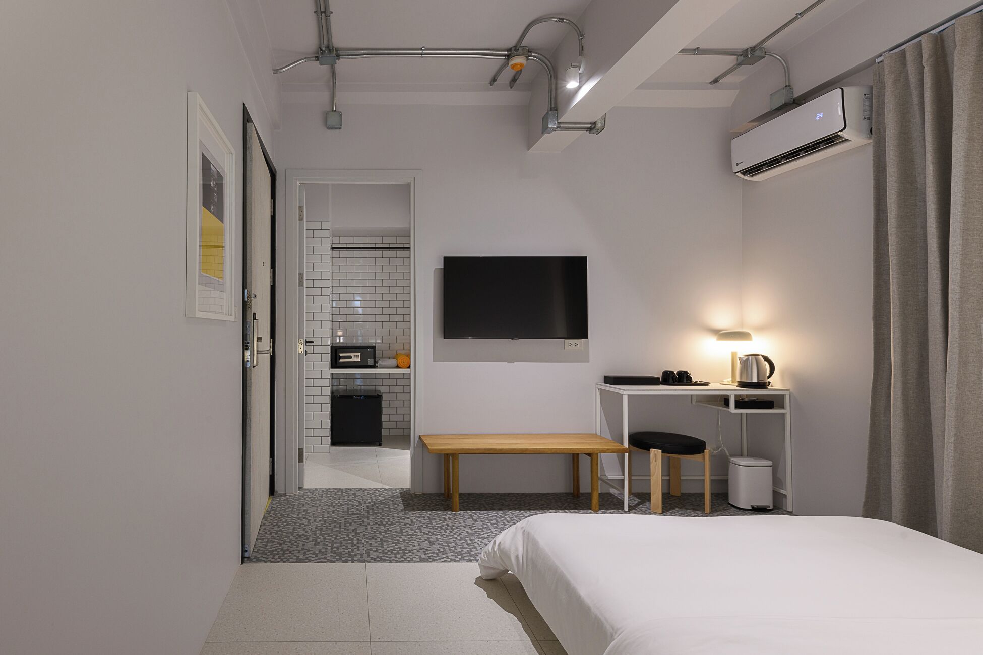 A modern hotel room with a bed, a desk with a lamp, a TV, and a kitchenette in the background.