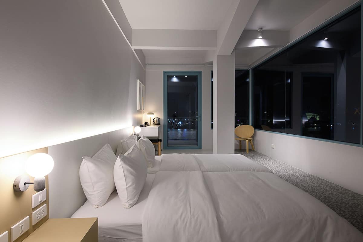 Quarto de hotel moderno com uma cama grande, luminárias laterais, uma mesa pequena e vista para a cidade à noite.