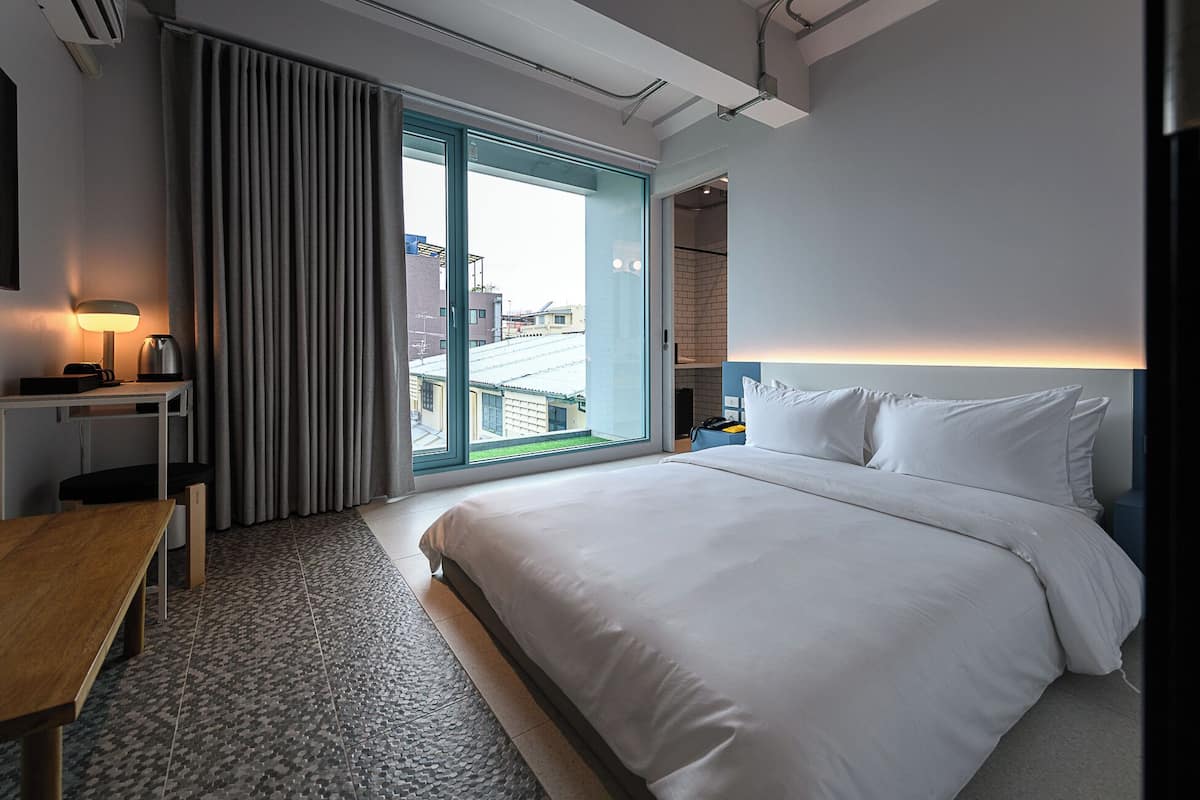 Quarto de hotel com uma cama grande, uma escrivaninha com luminária, vista através de uma porta de correr e um banheiro moderno visível através de uma porta aberta.