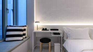 Habitación de hotel pequeña con una cama, un escritorio con lámpara, un taburete y un cesto de basura.