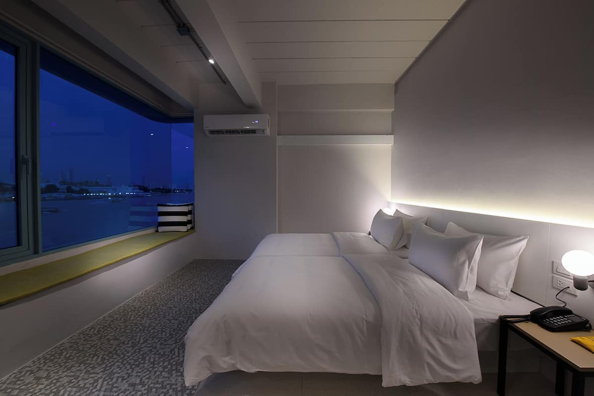 Quarto de hotel moderno com uma cama grande, abajur, mesa pequena com telefone e janela com vista para a paisagem urbana ao entardecer.