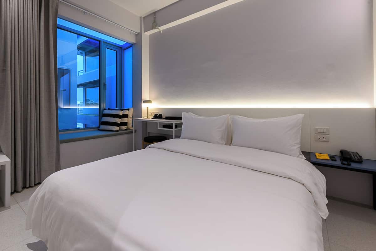 Quarto de hotel moderno com uma cama grande, uma mesa com luminária, uma mesinha de cabeceira com telefone e uma janela com vista para o exterior.