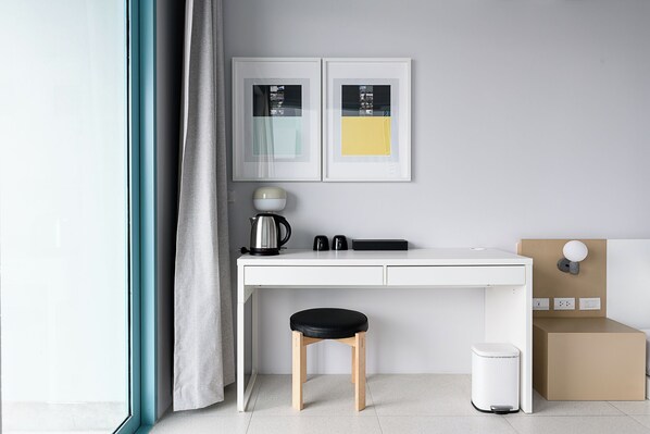 Habitación minimalista con un escritorio blanco, un taburete negro, un cesto de basura blanco y dos cuadros en la pared.