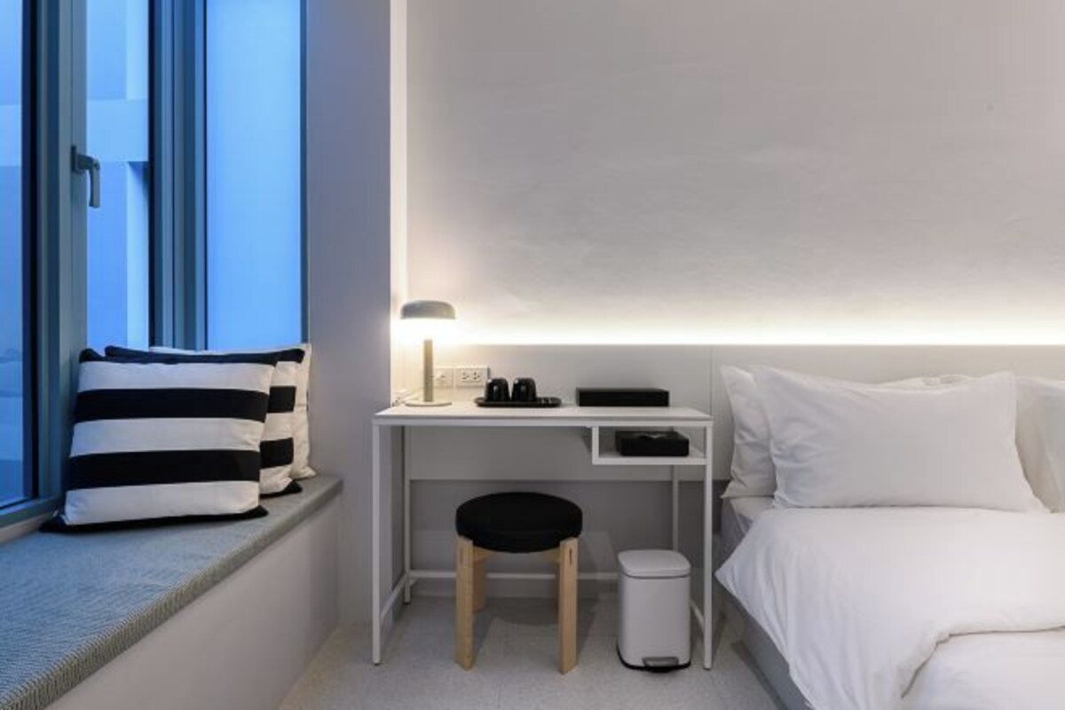 A hotel room with a bed, a desk with a lamp, a trash bin, and a window with pillows.