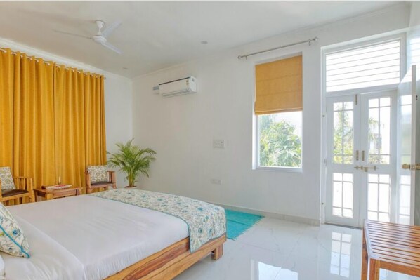 A bedroom with a bed, two chairs, a small table, a potted plant, and a window with a view of greenery. - Rohet Villa (Jaipur)