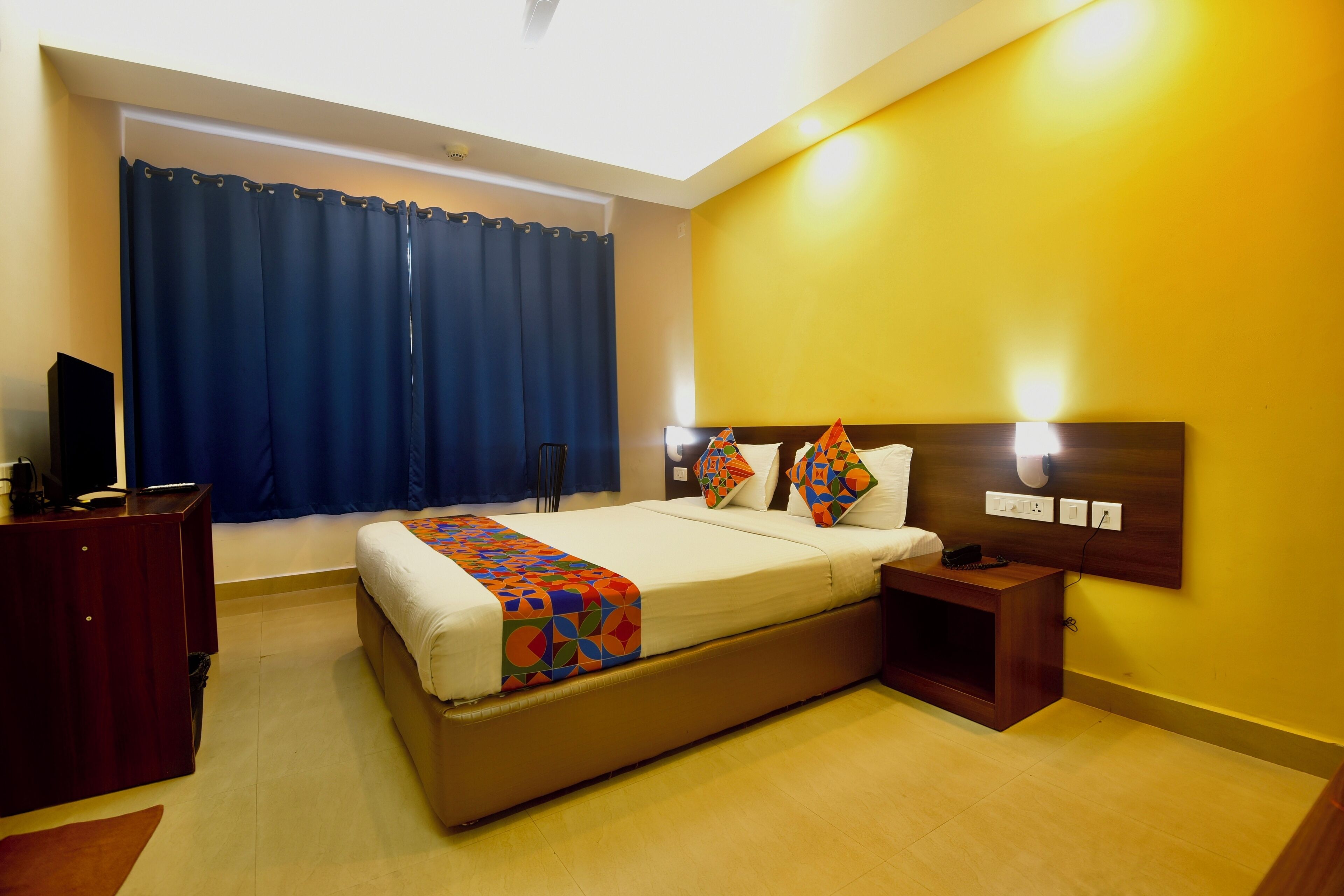 A hotel room with a bed, bedside tables, a desk, a chair, a TV, and a window with curtains.