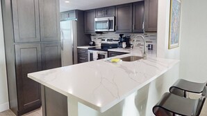 Uma cozinha moderna com armários escuros, uma ilha de mármore branco, eletrodomésticos de aço inoxidável e uma única cadeira.