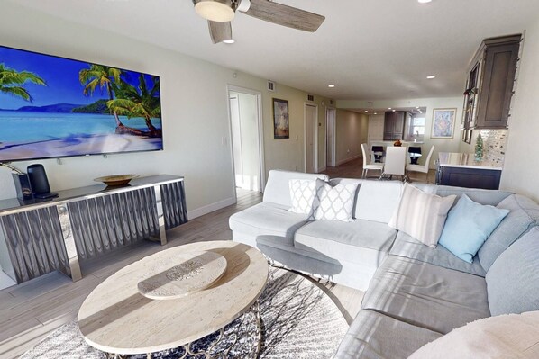 A modern living room with a large flat-screen TV, a white sofa, a round coffee table, and a dining area in the background.