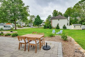 Un patio con una mesa y sillas de madera, un pequeño jardín con césped y algunos árboles, y una casa blanca al fondo.
