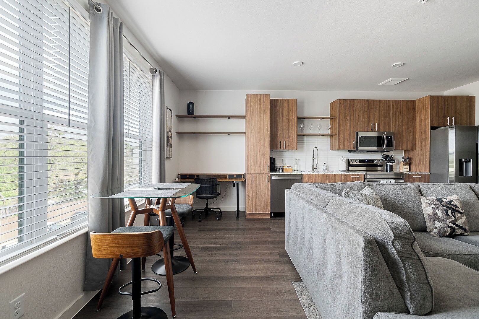Una cocina moderna con armarios de madera, una zona de comedor con mesa y sillas de vidrio, y un sofá gris con un cojín estampado.