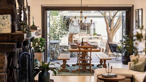 An interior with a dining area, a sofa, and a view through a glass door to an outdoor space with a table and chairs.