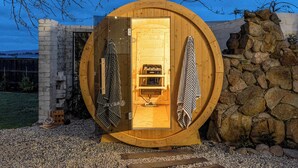 A wooden sauna with a circular door, a bench inside, and a stone wall in the background.