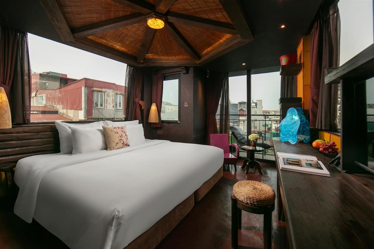 Une chambre d’hôtel avec un grand lit, une petite table avec une chaise, un balcon donnant sur des immeubles et un plafond en bois.