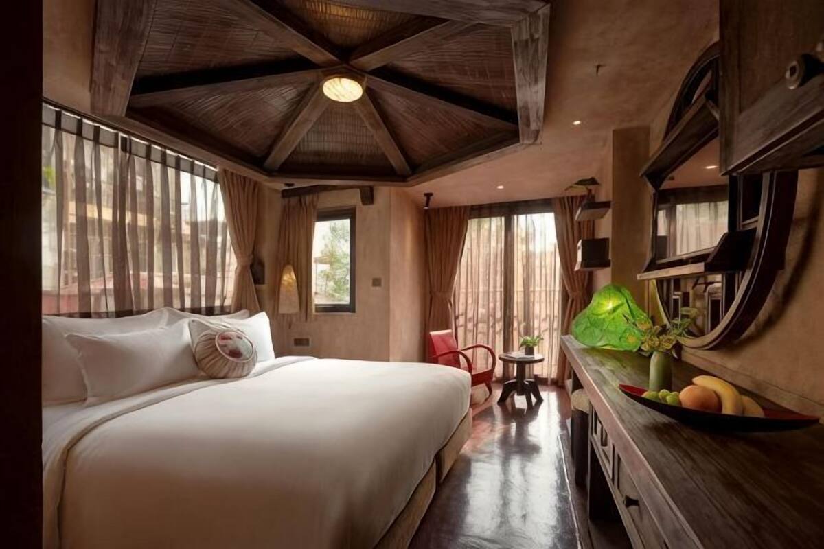 Une chambre d’hôtel avec un grand lit, un bureau en bois sur lequel se trouve un panier de fruits, et une petite table avec une chaise.